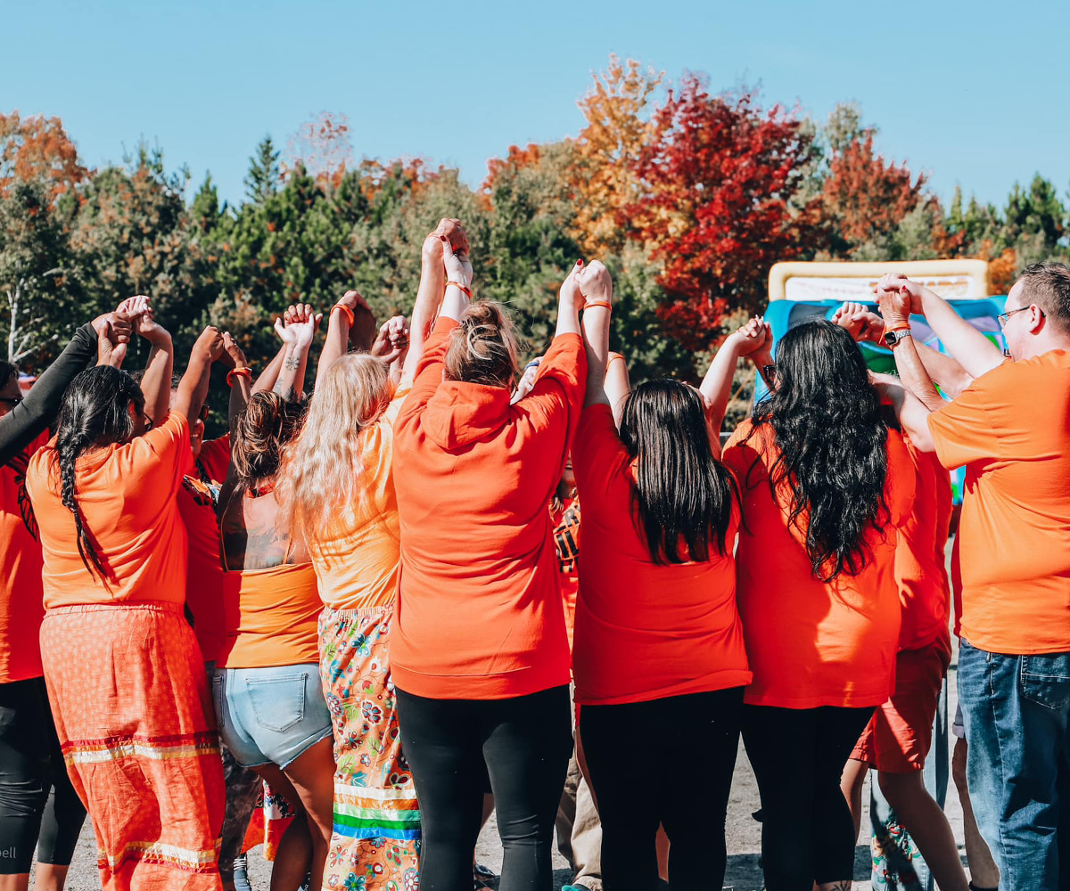 A group of people in orange shirts stand in a circle holding hands outdoors, with trees in the background.
