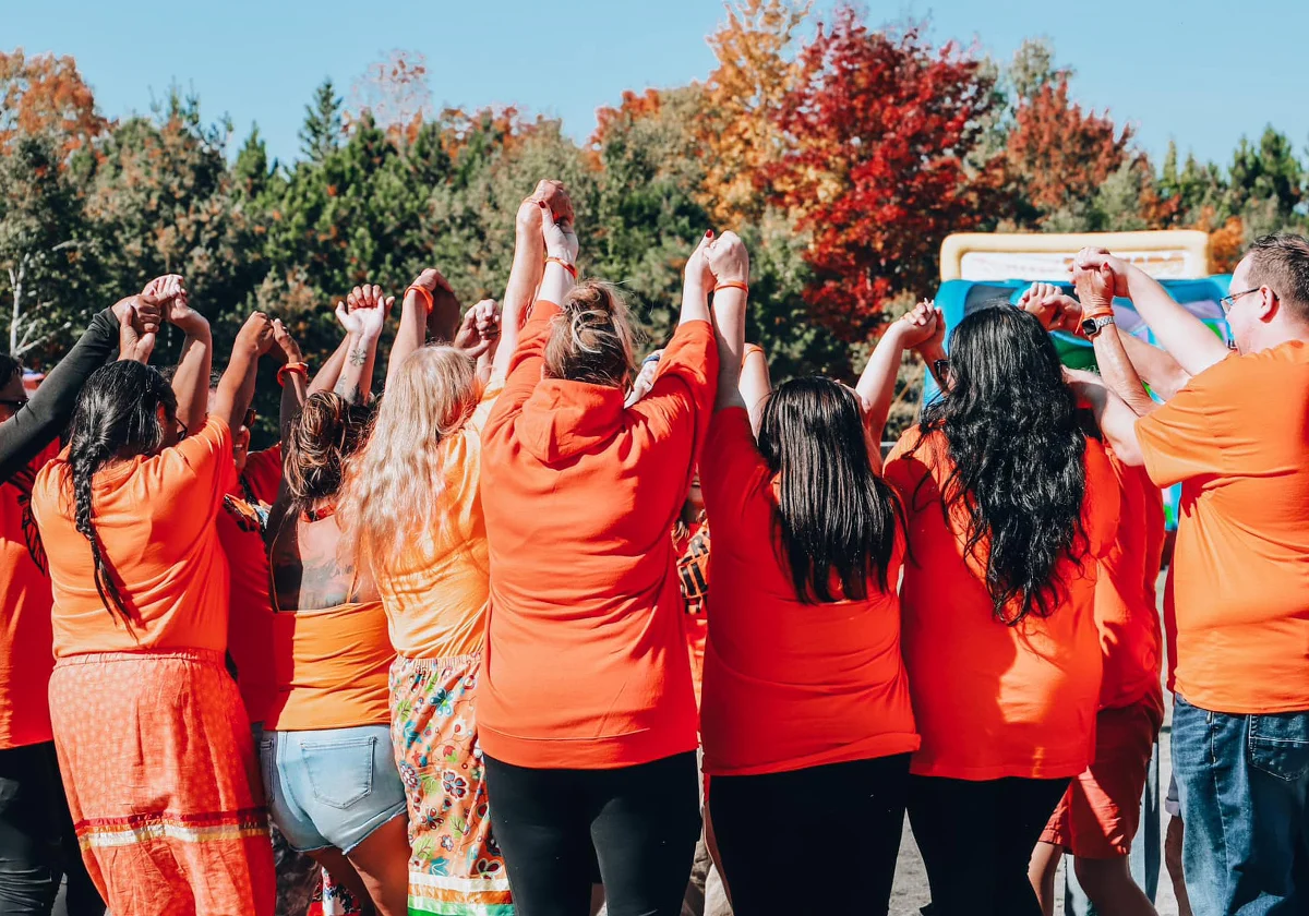 A group of people in orange shirts stand in a circle holding hands outdoors, with trees in the background.
