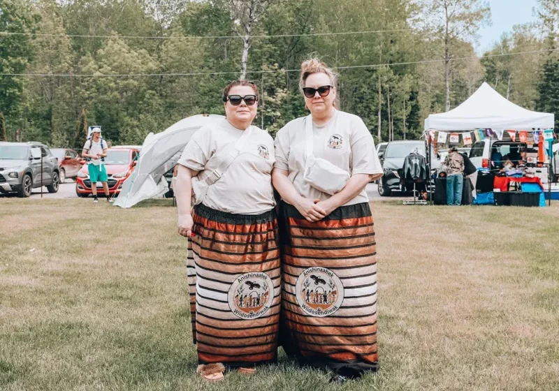 Two women in traditional attire stand on grass at an outdoor event with tents and cars in the background.