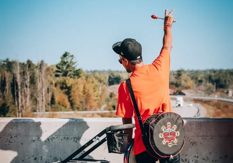 Person in orange shirt and black cap raises hand on an overpass, with a scenic road and trees in the background.