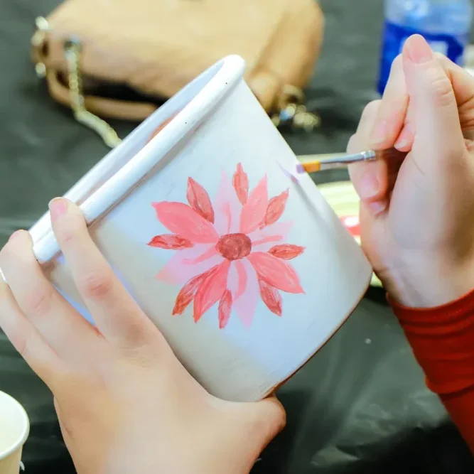 A person paints a red flower on a white flowerpot with a small brush.