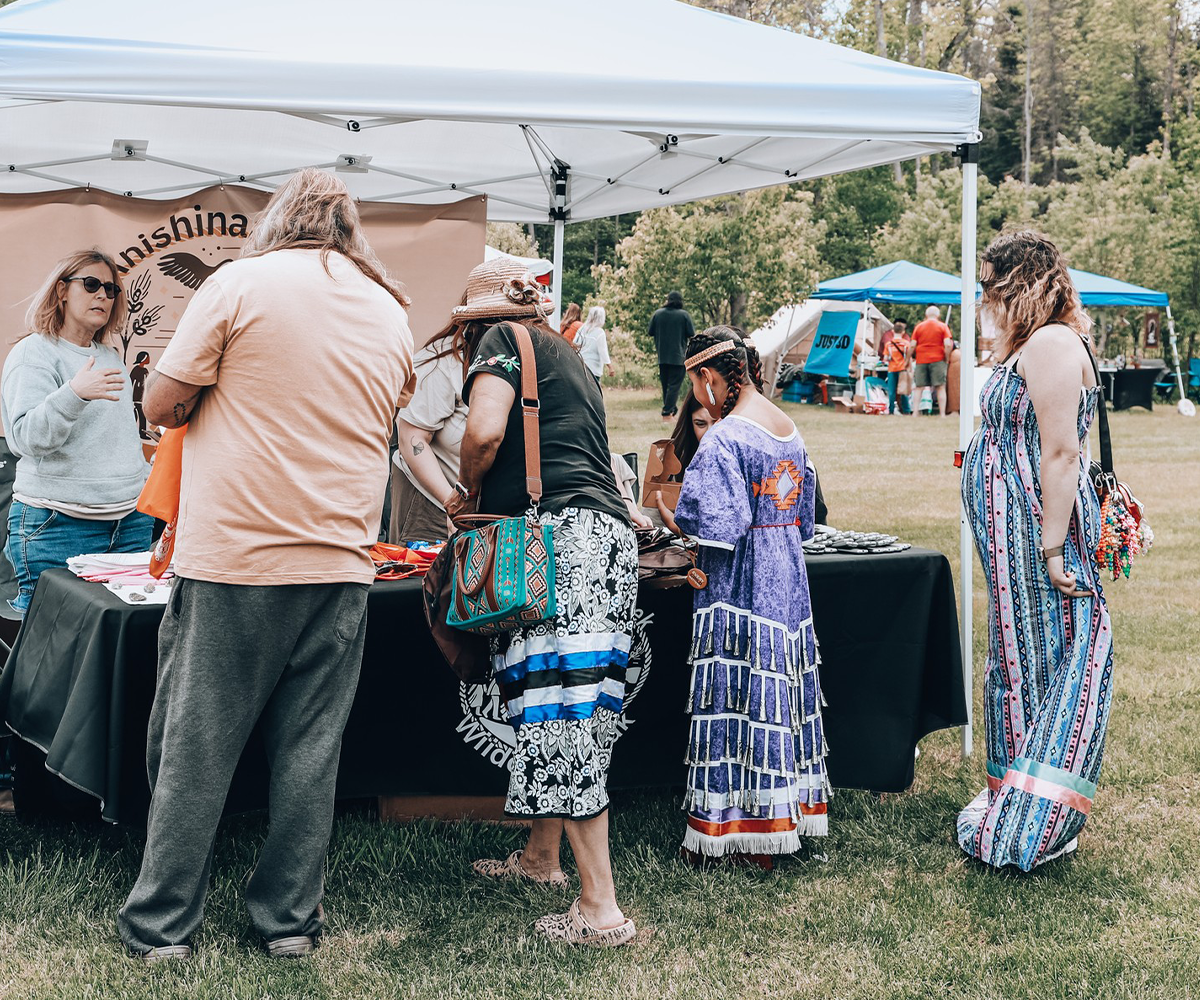 People browse items at an outdoor market booth, some wearing colorful, traditional, Indigenous clothing.