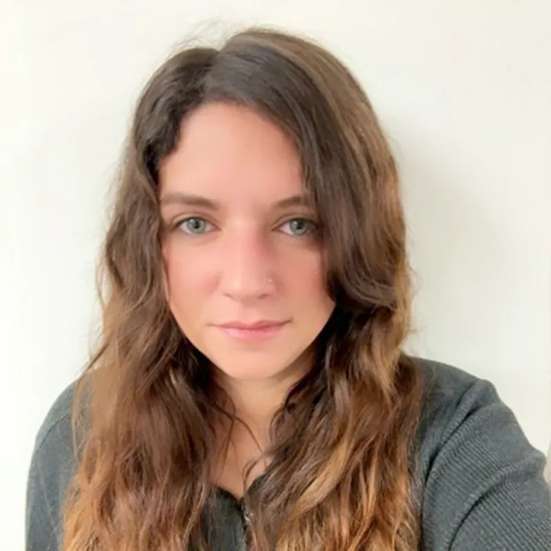 Woman with long wavy brown hair and green eyes, wearing a dark top, in front of a plain white background.