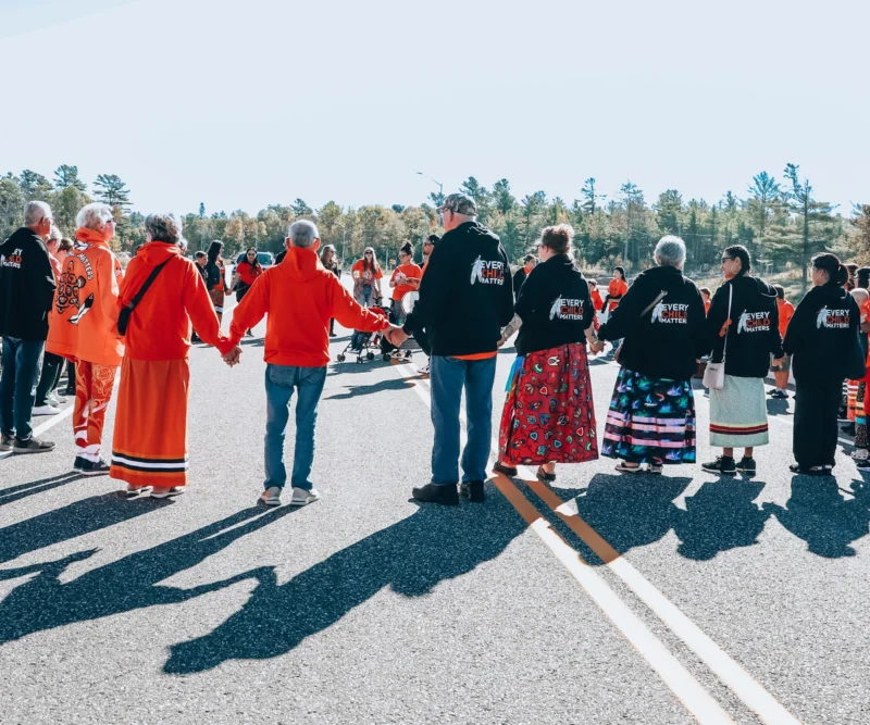 People wearing orange and black shirts stand in a circle, holding hands on a sunny road, with trees in background.