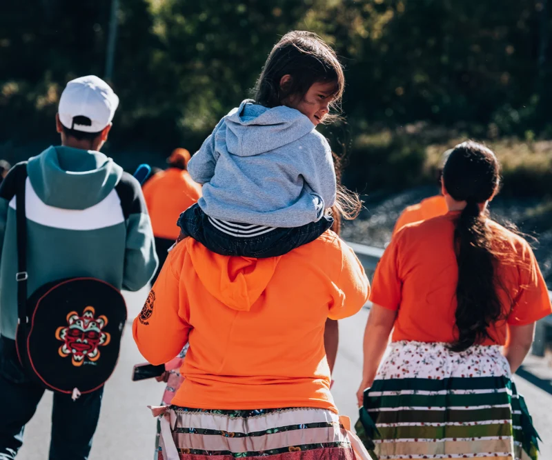 A child in a gray hoodie rides on an adult's shoulders among people wearing orange shirts outdoors.