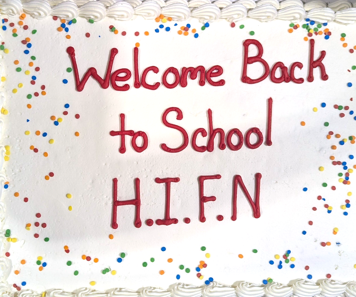 A white cake with "Welcome Back to School H.I.F.N" written in red icing and colorful sprinkles.