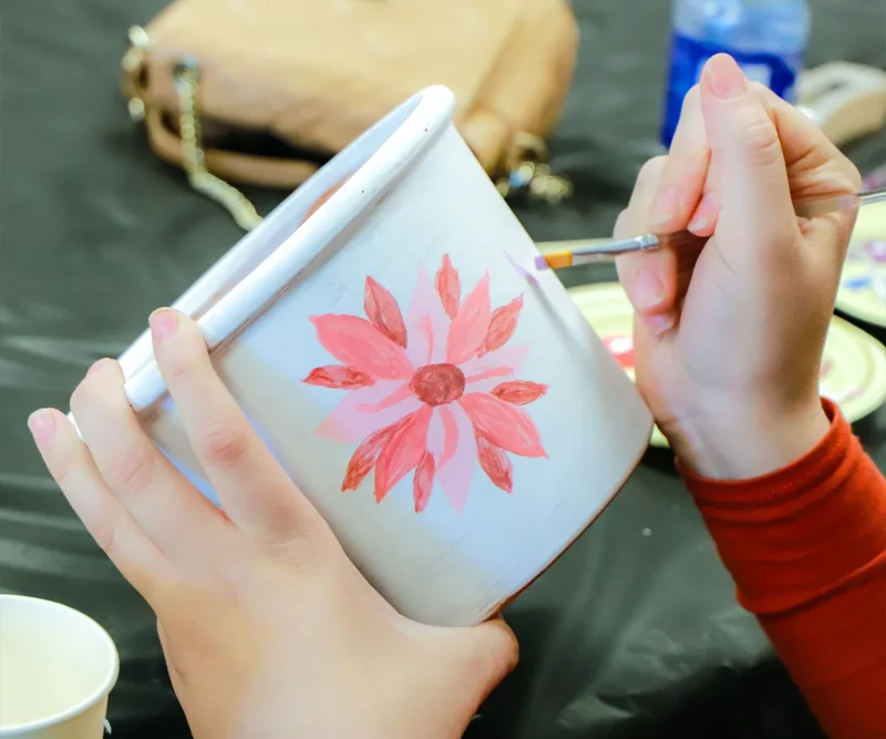 A person paints a red flower on a white flowerpot with a small brush.