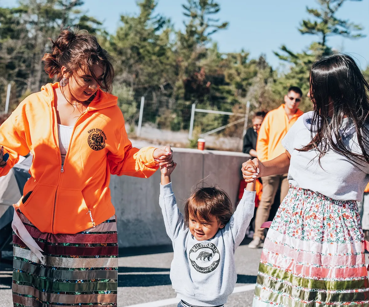 Two women in ribbon skirts hold hands with a smiling child outdoors on a sunny day.