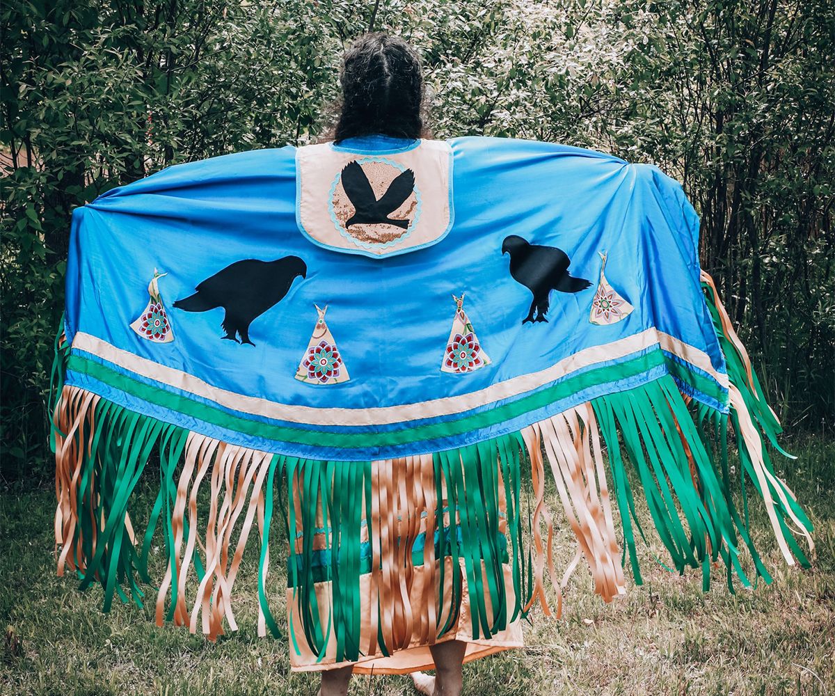 Person wearing a blue Indigenous shawl with bird and tipi designs, standing outdoors with arms extended.